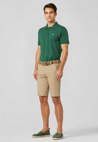 Groene poloshirt, beige shorts en groene espadrilles. Gebloemde riem. Poloshirt heeft een kraag en korte mouwen. Casual zomerse outfit.