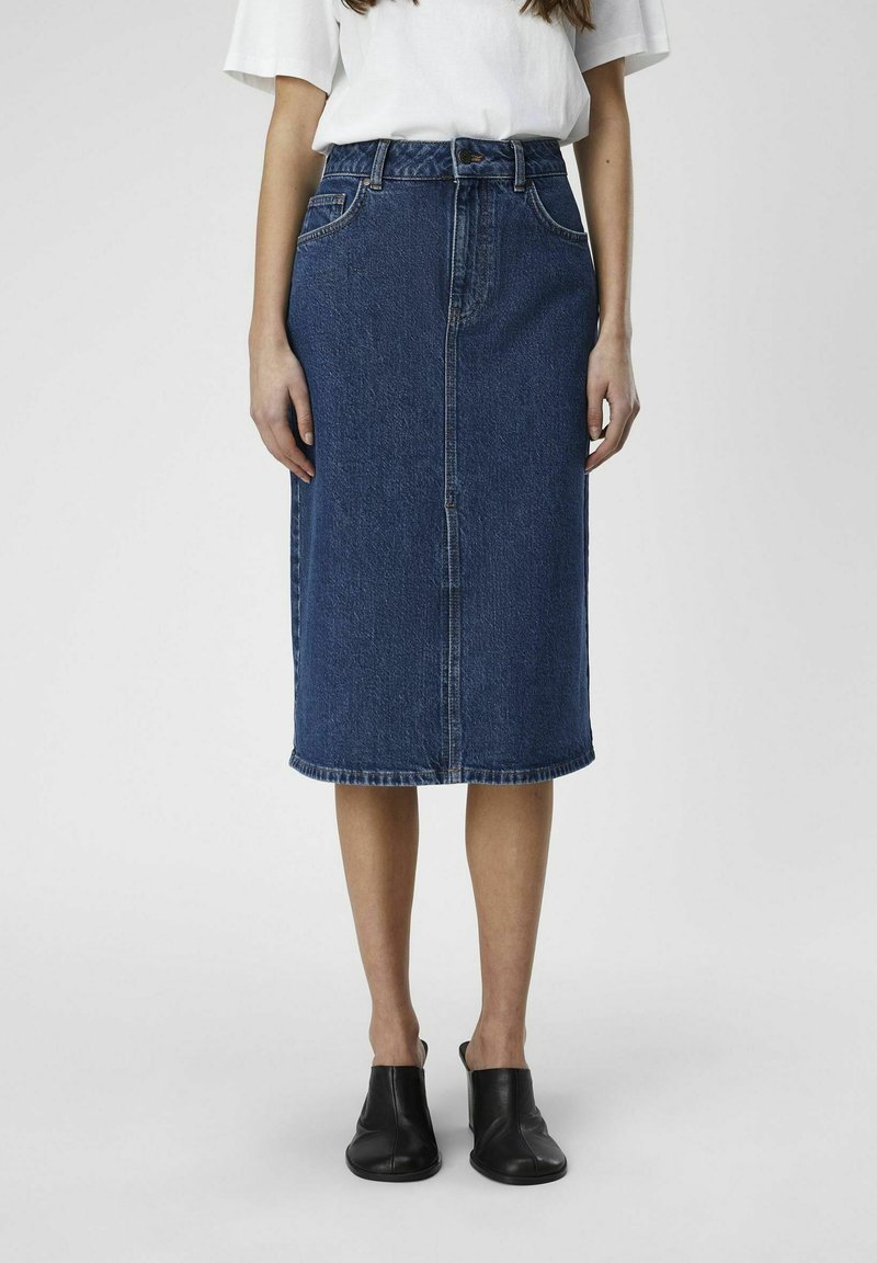 Denim midi-skjørt i blått med rett snitt, frontlukk med knapper, beltehemper og hemmede kanter; brukt med svarte slip-on-sko.