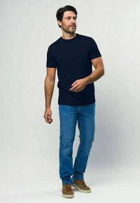 Tumši zils īsām piedurknēm T-krekls ar mazu sarkanu logotipu uz krūtīm, saskaņots ar gaiši ziliem džinsiem un brūnām zamšādām sporta apaviem.