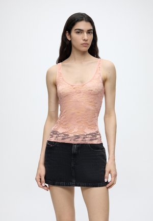Femme aux longs cheveux foncés porte un débardeur en dentelle rose clair et une mini-jupe en jean noire, debout sur un fond blanc uni.