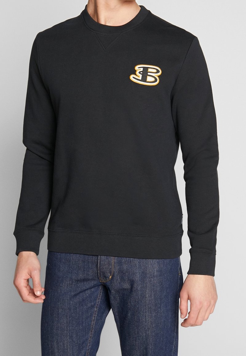Homme portant un sweat-shirt noir à manches longues avec un logo "B" jaune et blanc sur la poitrine, associé à un jean bleu, sur un fond uni.