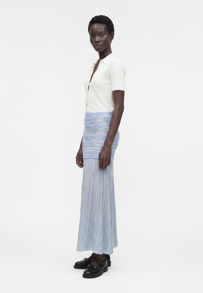 Γυναίκα με κοντά μαλλιά που φοράει λευκό κοντομάνικο τοπ με κουμπιά, μπλε πλεκτή μακριά φούστα και μαύρα μοκασίνια, όρθια πλαγίως.