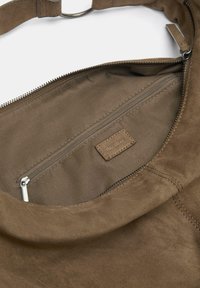 Brun mockaväska insida med en dragkedjeficka, mjuk textur och en etikett med präglad logotyp. Metallkedjedragkedjans detaljer synliga.