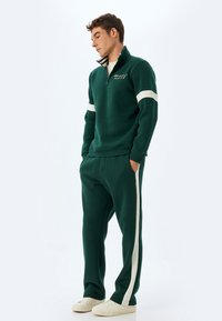 Dunkelgrüner Fleece-Trainingsanzug mit Reißverschlusskragen, weißen Streifen an den Ärmeln und Seiten der Hose, lässiger Schnitt, gerippte Bündchen und seitlichen Taschen.