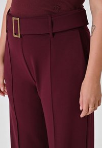 Pantalon bordeaux avec une taille large, doté d'une boucle en or rectangulaire. Le tissu est lisse avec des plis latéraux subtils.