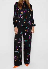 Černý outfit s patterned halenkou a širokými kalhotami s geometrickými tvary v purpurové, oranžové a tyrkysové barvě. Zahrnuje oranžové podpatky.