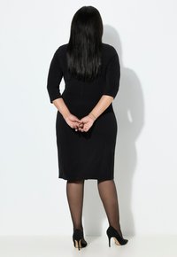 Vestido negro con mangas tres cuartos, silueta ajustada y bajo a la altura de la rodilla. Llevado con tacones altos negros y medias opacas.
