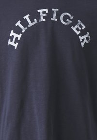 Camiseta azul marino con el texto "HILFIGER" en blanco desvaído en un diseño arqueado, fabricada con un suave tejido de algodón de textura lisa.