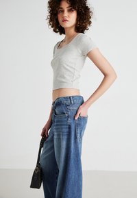 Grå kort topp med korte ermer, parret med vide blå denimjeans. Modellen holder en liten svart håndveske med gull detaljer. Minimalistisk bakgrunn.