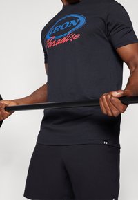 Atletyczna osoba trzymająca czarny sztangę, ubrana w czarną koszulkę "Iron Paradise" i czarne szorty, na jednolitym tle.