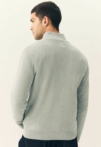 Hombre de perfil izquierdo vistiendo un suéter texturizado de cuello alto gris claro y pantalones oscuros frente a un fondo liso.