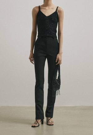 Schwarzes, mit Perlen besetztes Tanktop mit dünnen Trägern, eng anliegende schwarze Hose und Riemchenabsätze. Kleine, schwarze Fransenhandtasche in der Hand.