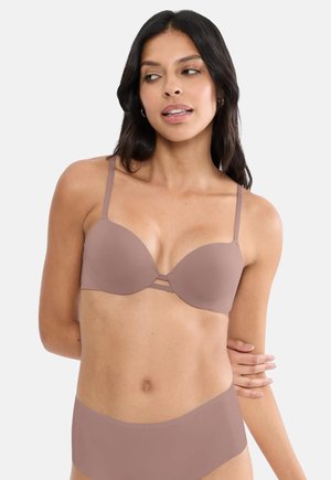 Γυναίκα που φοράει μοβ αόρατο σουτιέν και εσώρουχα με ψηλή μέση, ποζάροντας με το ένα χέρι πάνω στο σώμα της, κοιτάζοντας προς το πλάι, σε λευκό φόντο.