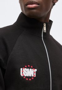 Κοντινό πλάνο ατόμου που φοράει μαύρο σακάκι με φερμουάρ με το λογότυπο USMNT περιτριγυρισμένο από κόκκινα αστέρια στο αριστερό στήθος.