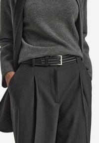 Person som bär en mörkgrå stickad tröja instoppad i svarta veckade byxor, med händerna i fickorna och ett svart vävt bälte med silverspänne.