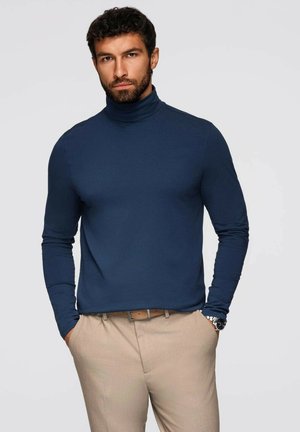 Marineblauwe coltrui van gladde stof met lange mouwen, gecombineerd met beige broek en een bruine riem. Eenvoudig, aansluitend design.