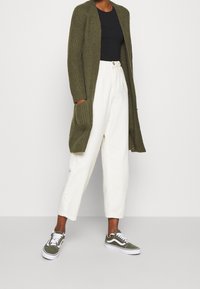 Oliivinvihreä ribatty neuletakki etutaskuilla, musta istuva toppi, kermaväriset leveät housut ja vihreät lenkkarit valkoisilla yksityiskohdilla.