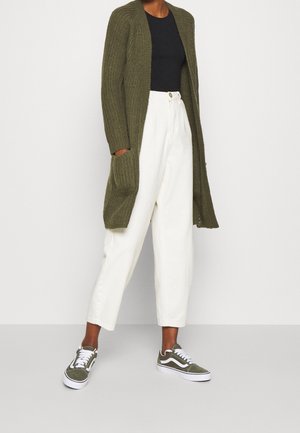 Olijfgroen ribgebreid vest met voorzakken, zwart aansluitend topje, crèmekleurige wide-leg broek en groene sneakers met witte accenten.