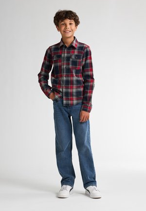 Camisa de franela a cuadros rojos y navy con dos bolsillos en el pecho, combinada con jeans azules y zapatillas blancas. Cabello rizado y fondo neutro.