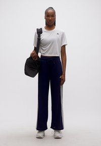 Γυναίκα που στέκεται φορώντας λευκό crop top Adidas, μπλε σκούρο παντελόνι φόρμας Adidas, λευκά αθλητικά παπούτσια και κρατώντας μια μαύρη τσάντα ώμου.