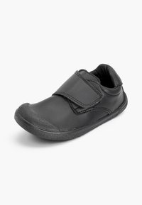 Chaussure noire pour enfant avec une large bride Velcro unique et une semelle en caoutchouc texturée, présentée légèrement inclinée sur un fond uni.