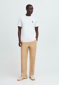 Hvit bomull T-skjorte med en liten svart logo, sammen med lysebeige rette bukser og beige sneakers med brune detaljer.