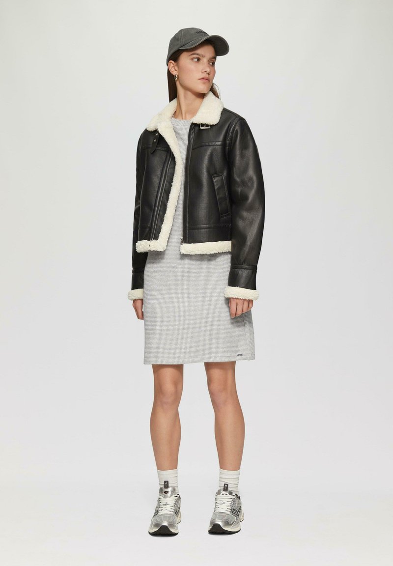 Schwarze Lederjacke mit weißem Schafslederfutter und -bündchen, kombiniert mit einem grauen gemusterten Kleid und silbernen Sneakers. Eine Mütze vervollständigt den Look.