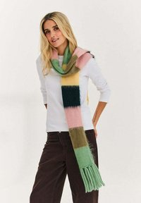 Kobieta o długich blond włosach ubrana w białą bluzkę z długim rękawem, ciemne spodnie i długi, wielokolorowy szalik w paski z frędzlami, stoi na tle jednolitego tła.