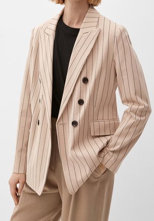 Blazer croisé beige à fines rayures avec boutons noirs, porté sur un haut noir, assorti à un pantalon beige.