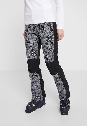 Person trägt schwarz-weiße gemusterte Skihose und schwarze Skischuhe, steht vor einem schlichten hellen Hintergrund.