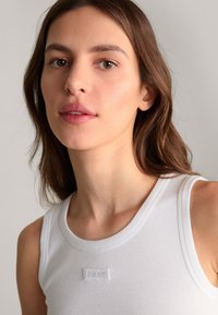 Top tank bianco a coste con scollatura rotonda e una piccola etichetta con logo sul petto. Il tessuto appare morbido ed elastico.