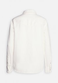 Chemise blanche à manches longues, boutonnée avec col, coutures visibles au niveau de l'empiècement arrière, et poignets boutonnés.