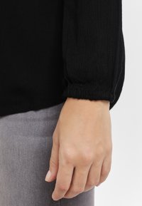 Svart långärmad topp med ribbad textur och elastiska manschetter; hand vilar på ett grått tyg, vilket framhäver ärmdetaljen.