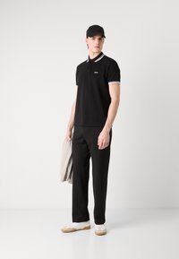 Schwarzes Poloshirt mit kontrastierendem weiß-blau gestreiften Kragen, kombiniert mit schwarzen Hosen, hält einen hellgrauen Pullover und trägt weiße Sneakers.