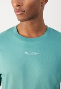 Zöldeskék pamut póló kerek nyakkal, fehér nyomtatott "Marc O’Polo" és "EST. IN STOCKHOLM" logóval a mellkasán. Simább textúra.