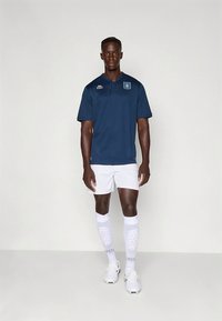 Tummansininen lyhythihainen poolopaita, jossa kaulus, valkoiset shortsit, kuvioidut valkoiset sukat ja valkoiset urheilukengät. Koristeena logoyksityiskohtia.