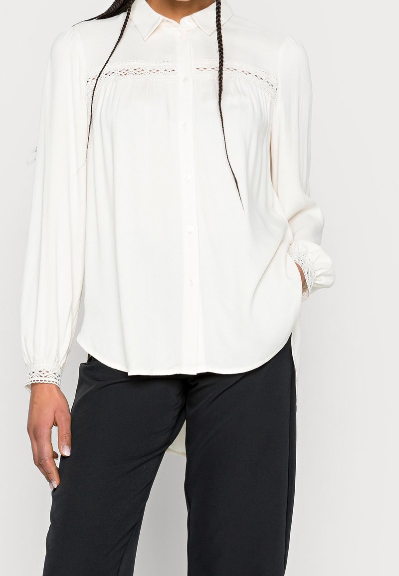 Vit blus med knäppning framtill, spetsdetaljer på axlarna, långa ärmar med elastiska muddar och rundad nederkant. Stylt med svarta byxor.