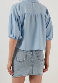 Blusa azul clara de manga corta con la parte trasera fruncida y puños elásticos, combinada con una falda de mezclilla ajustada en un lavado claro.