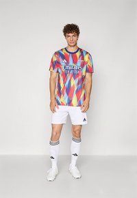 Barevný dres Adidas v abstraktním vzoru červené, modré a žluté barvy, s černým límcem a logem "Emirates". Kombinován s bílými šortkami a ponožkami.