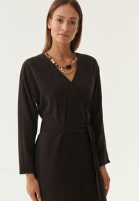Schwarzes Wickelkleid mit langen Ärmeln und Taillenknoten, aus glattem Stoff gefertigt. Akzentuiert mit einer Statement-Halskette, die goldene und schwarze Elemente enthält.