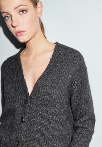 Donkergrijze gebreide cardigan met een diepe V-hals, gestructureerde stof en vier zwarte knopen aan de voorkant. Getailleerde pasvorm met lange mouwen.
