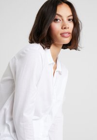 Camicia bianca con bottoni dal taglio rilassato e tessuto morbido, con colletto classico e dettagli con bottoni discreti, indossata da un modello.