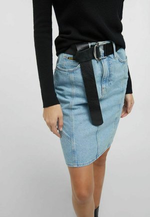 Frau trägt ein schwarzes langärmeliges Oberteil, das in einen hellblauen Jeansrock mit locker hängendem schwarzem Gürtel gesteckt ist.