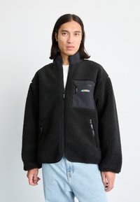 Schwarze Fleecejacke mit Reißverschluss vorne, hohem Kragen und strukturierter Oberfläche. Verfügt über eine Brusttasche mit einem farbigen Logo und seitlichen Reißverschlusstaschen.