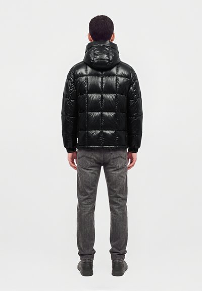 Homme portant une doudoune matelassée noire avec capuche, un jean gris et des chaussures noires, debout de dos sur un fond blanc uni.