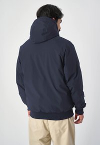 Dunkelblaues Kapuzenjacke mit glatter Textur und elastischen Bündchen, mit lässigem Schnitt und geripptem Taillenbund, von hinten betrachtet.