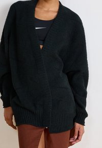 Person som bär en svart texturerad kofta över en svart sportbehå och en brun kjol med slits fram, poserande mot en ljus vägg.