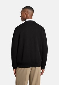 Schwarzer Strickpullover mit gerippten Bündchen und Saum, der eine strukturierte Oberfläche und überschnittene Schultern aufweist, getragen über einem weißen Hemd mit Kragen.