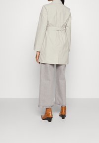 Manteau beige ceinturé à la coupe structurée, associé à un pantalon gris clair à jambes larges et des bottines marron à talons blocs.