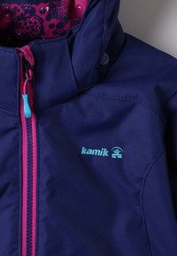 Námořnická voděodolná bunda s růžovými akcenty. Má vysoký límec, zipové zapínání s tyrkysovým táhlem a vzor uvnitř kapuce. Logo Kamik je viditelné.